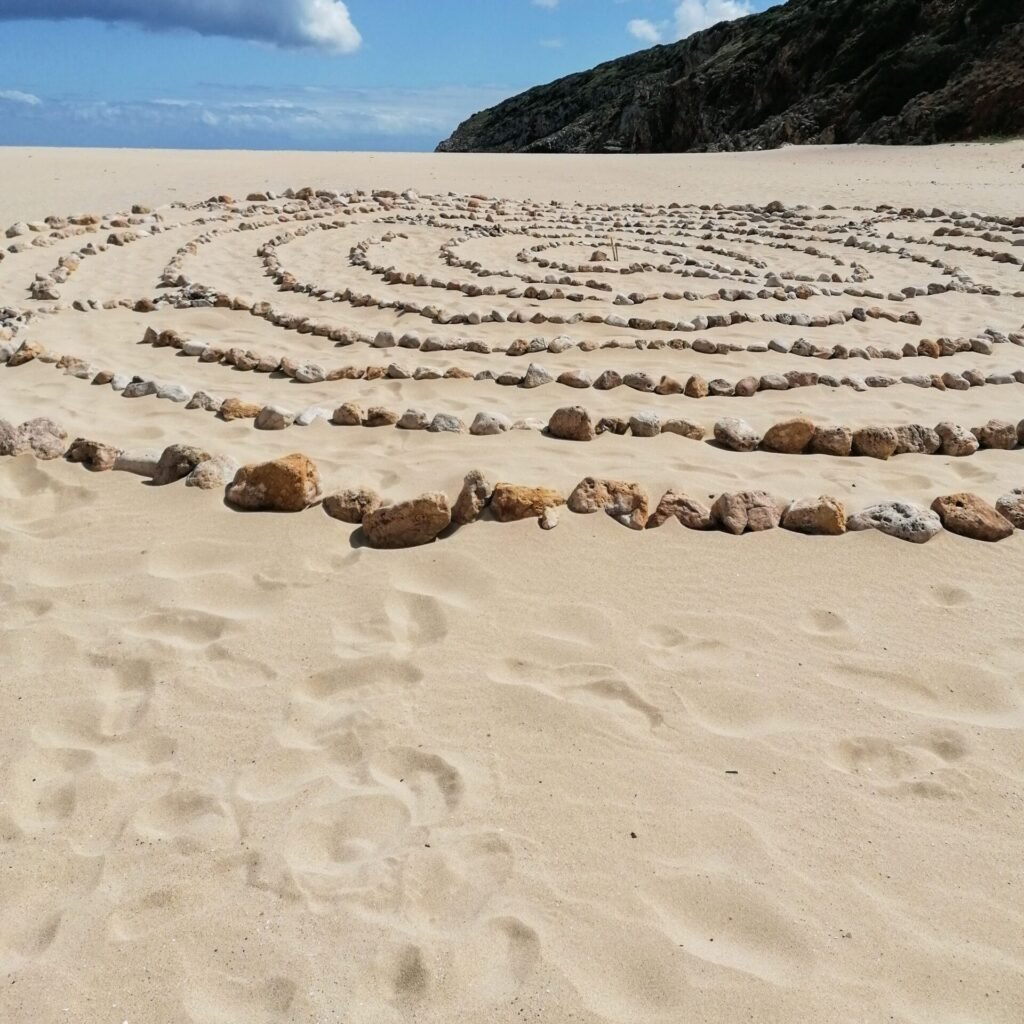 Artistic spiral stone design on a scenic sandy beach in Lisboa, Portugal under a partly cloudy sky.